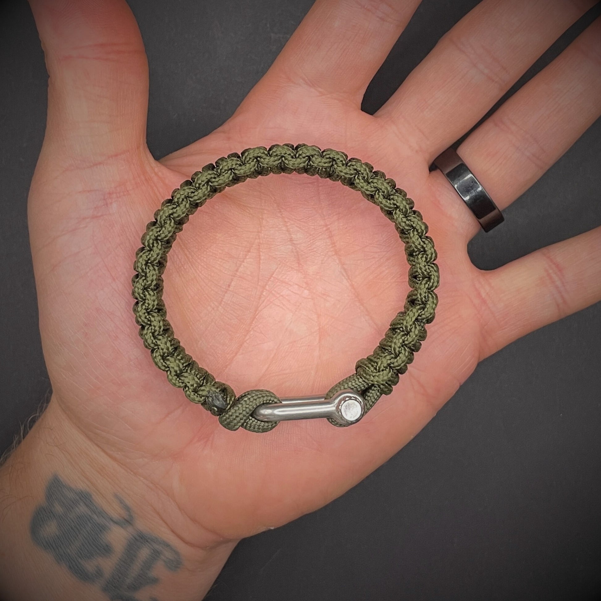 An army green paracord rope bracelet arranged on a dark grey surface.