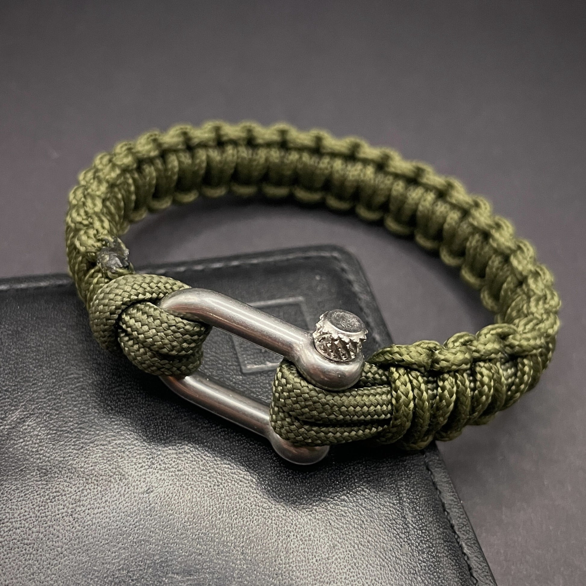 An army green paracord rope bracelet arranged on a dark grey surface.