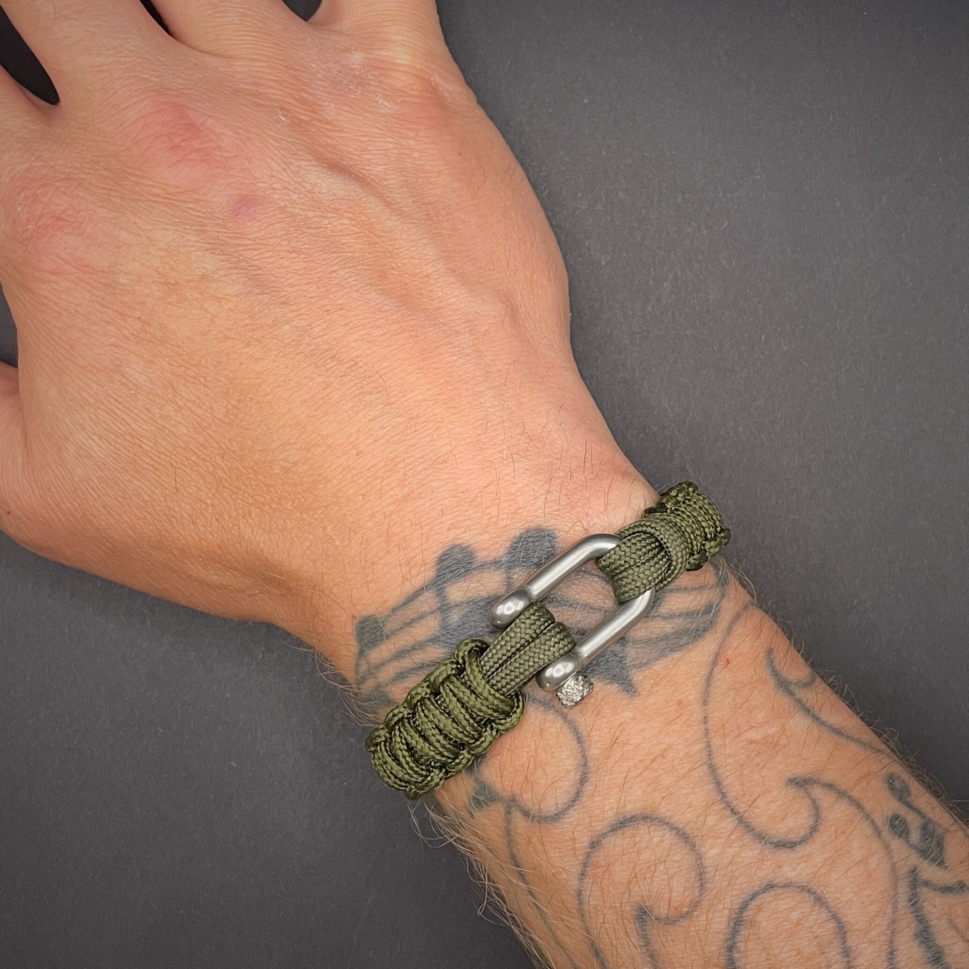 An army green paracord rope bracelet arranged on a dark grey surface with stainless steel shackle on a tattooed wrist.