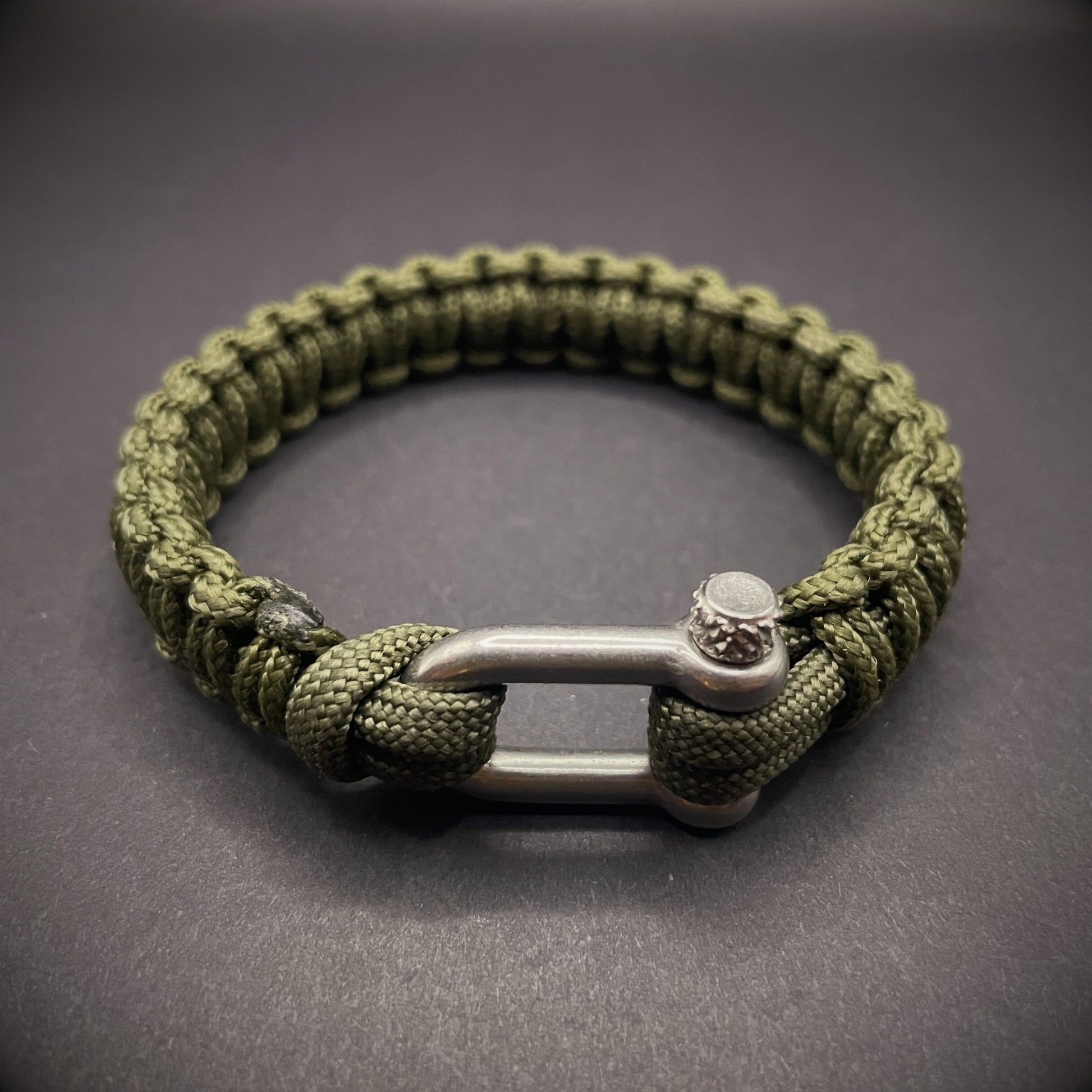 An army green paracord rope bracelet arranged on a dark grey surface.