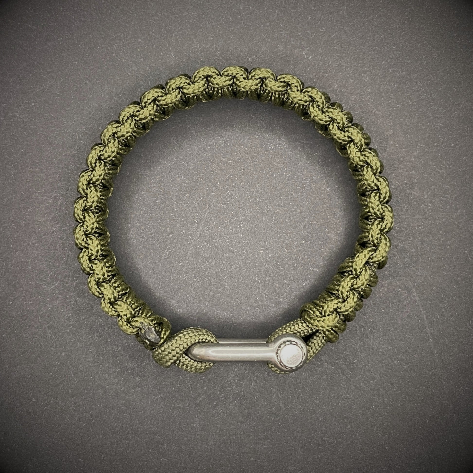 An army green paracord rope bracelet arranged on a dark grey surface.
