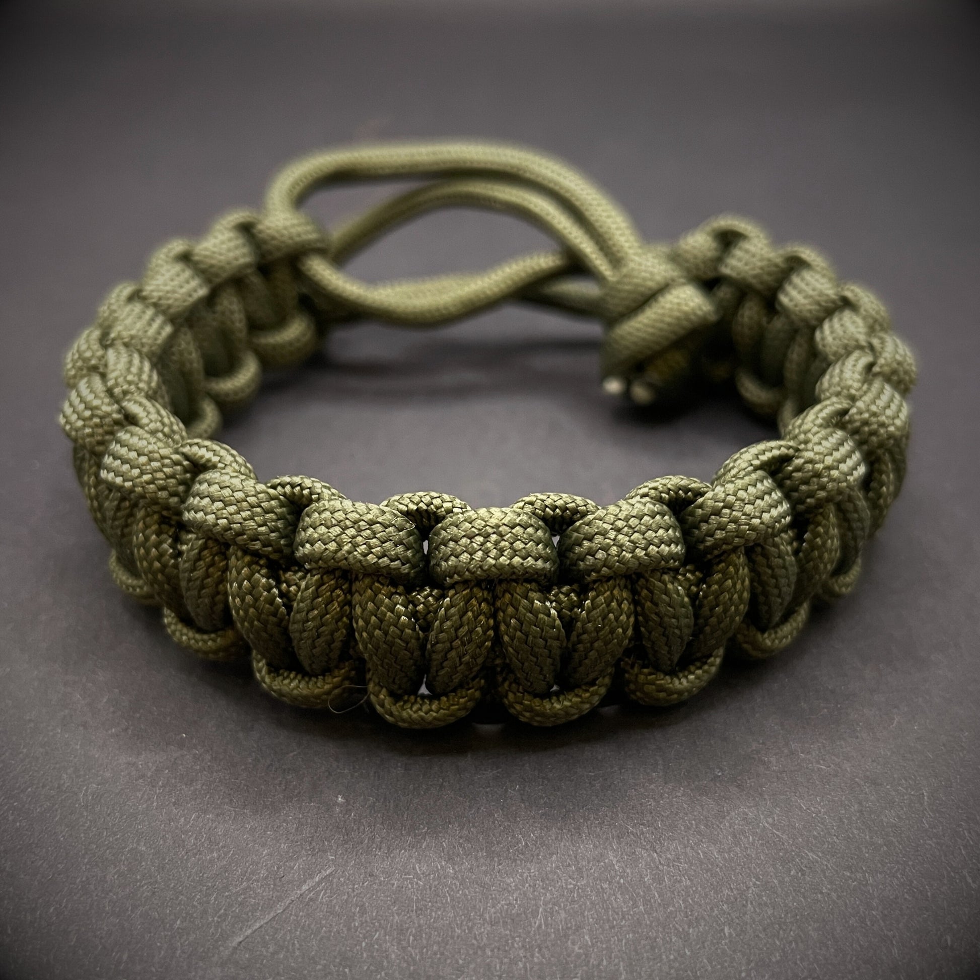 An Army green 550 paracord cobra knot bracelet displayed on a dark grey surface.